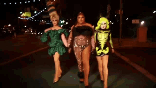 three drag queens are walking down the street at night holding hands