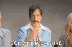 a man with a mustache is sitting in front of a group of people and says `` bon anniversaire '' .