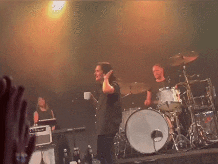 a man holding a cup stands on a stage with a drum set behind him