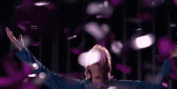 a woman is standing on a stage with her arms outstretched and purple confetti falling around her .