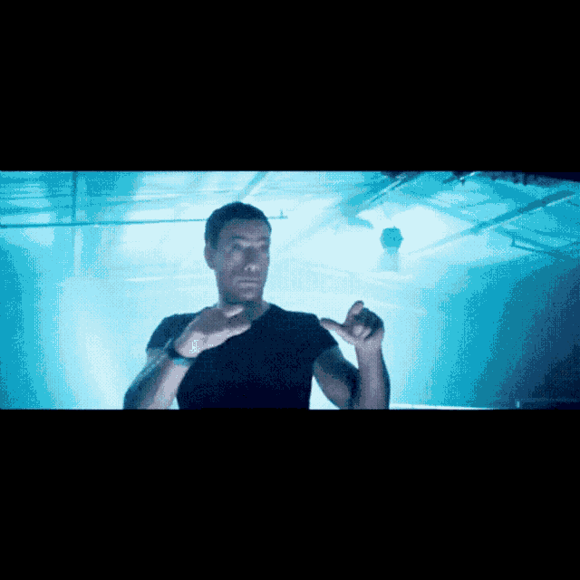a man in a black shirt stands in a dark room with a blue light behind him