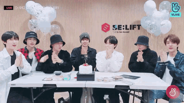 a group of young men are sitting around a table with a cake and balloons .