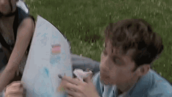 a man is sitting in the grass looking at a piece of paper with a drawing on it .