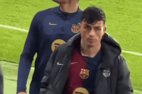 two men are walking on a soccer field . one of the men is wearing a jacket .