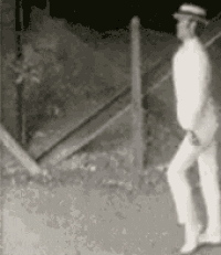 a black and white photo of a man in a suit and hat standing next to a fence at night .