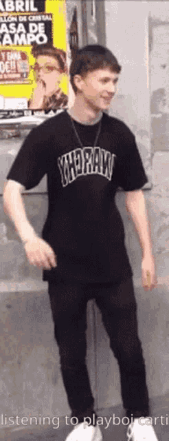 a young man wearing a black t-shirt that says yhdraw is standing in front of a wall .