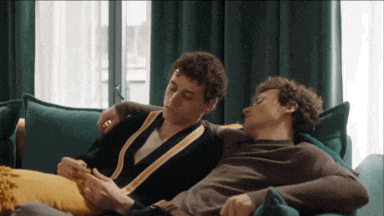 two men are laying on a couch and one of them is looking at his phone