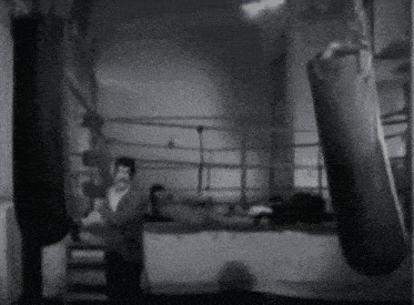 a black and white photo of a man in a boxing ring with punching bags hanging from the ceiling