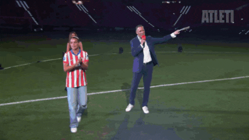 a man in a red and white atlético jersey stands on a soccer field with his arms in the air