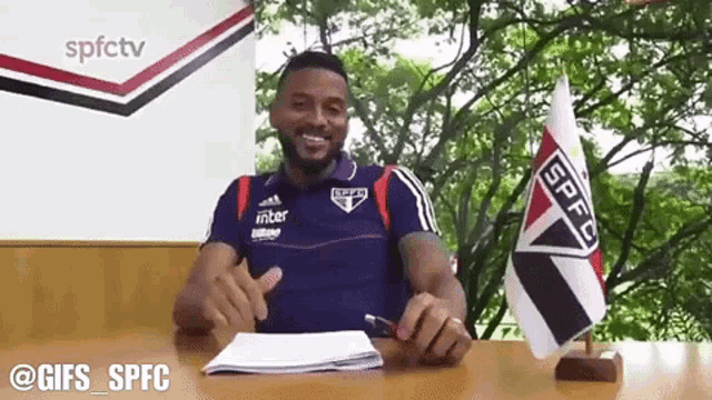 a man is sitting at a desk with a spfc flag in front of him