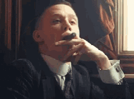 a man in a suit is smoking a cigar in front of a window .