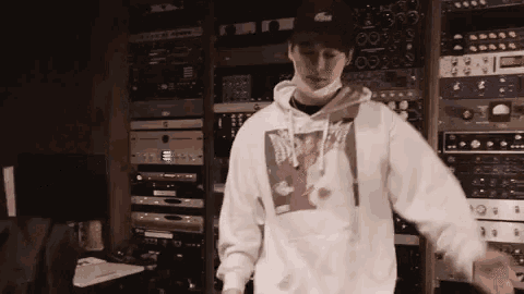 a man wearing a white hoodie and a black hat is standing in front of a rack of equipment in a recording studio .