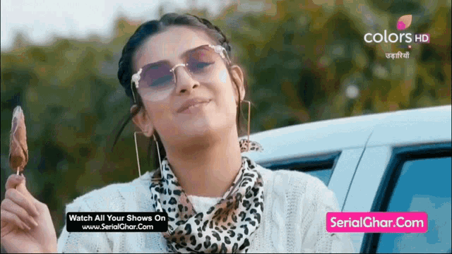 a woman wearing sunglasses and a scarf is holding an ice cream cone in front of a car .
