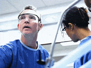 a man wearing goggles talks to a woman in a blue scrub