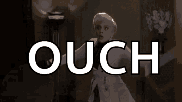 a woman in a white coat is standing in a room with the word ouch written on the screen .