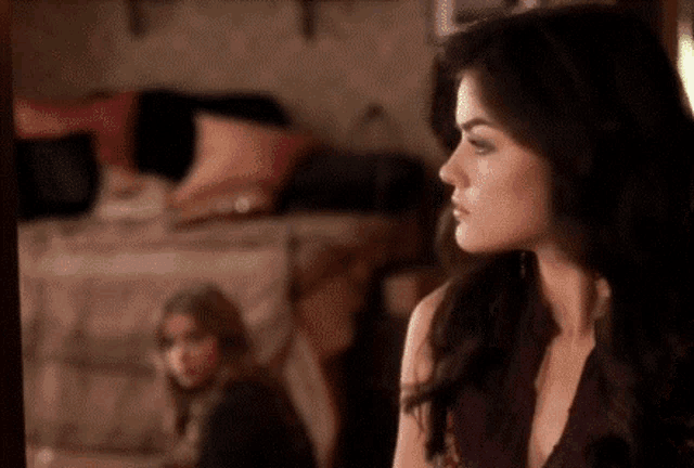 a woman is looking at herself in a mirror while another woman sits on a couch in the background .