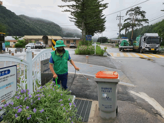 a man in a green vest spraying purple flowers next to a trash can that says easywood