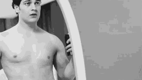 a shirtless man is looking at himself in a mirror while holding a spray bottle .