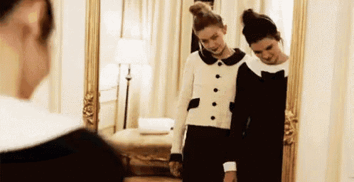 two women are standing next to each other in front of a mirror and looking at themselves .