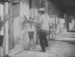 a black and white photo of a man in a hat walking down a street .