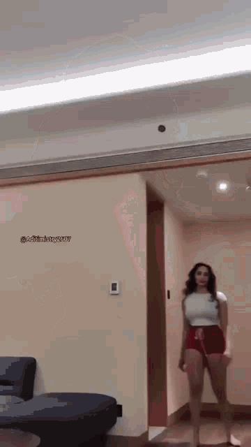 a woman in a white top and red shorts is standing in a room next to a couch .