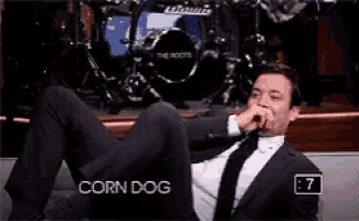a man in a suit and tie is sitting on a couch with his legs crossed and covering his nose with his hand .