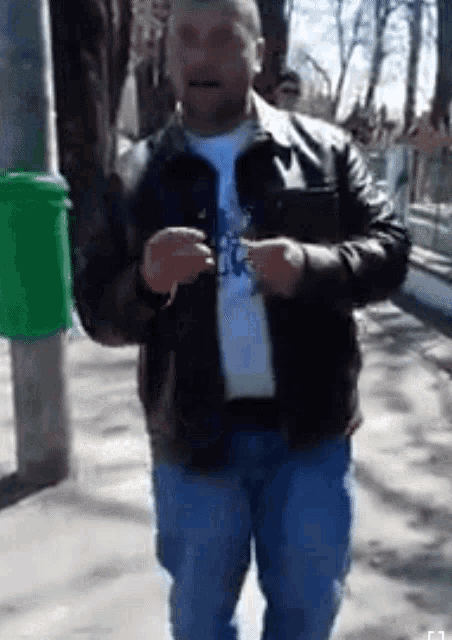 a man in a leather jacket is standing in front of a green trash can ..