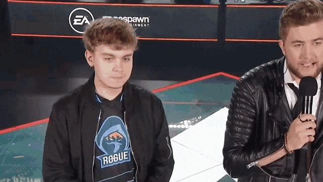 a man wearing a rogue shirt stands next to another man with a microphone