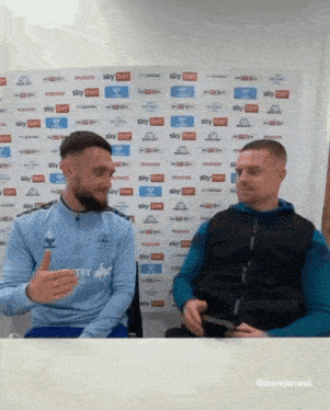 two men are sitting in front of a wall that has sky bet written on it