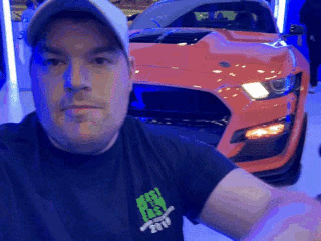a man wearing a black shirt that says ' mustang ' on it stands in front of an orange mustang