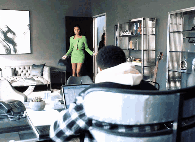 a woman in a green dress stands in a living room next to a man sitting in a chair