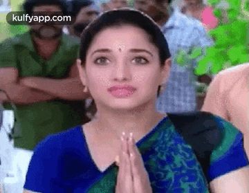 a woman in a blue shirt is standing in front of a crowd with her hands folded in front of her face .