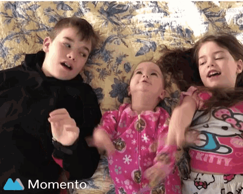 a boy and two girls are laying on a bed with a momento logo on the bottom