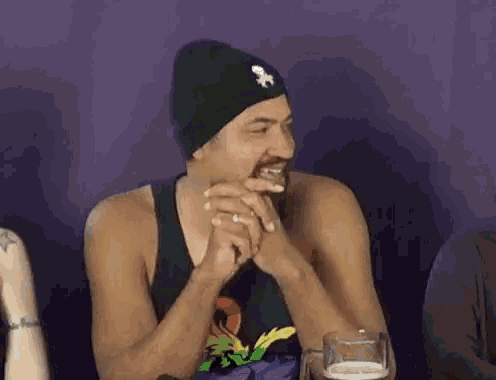 a man wearing a beanie is laughing while sitting at a table with a glass of beer in front of him .