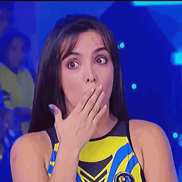 a woman is covering her mouth with her hand while wearing a yellow and blue tank top .