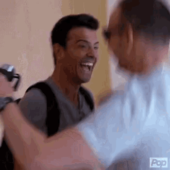 two men are hugging each other and laughing . one of the men is wearing a backpack .