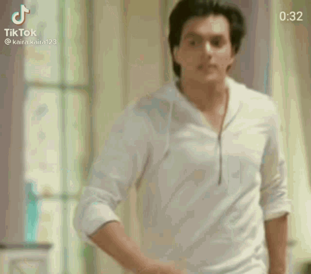 a man in a white shirt is standing in front of a window with his hands in his pockets .