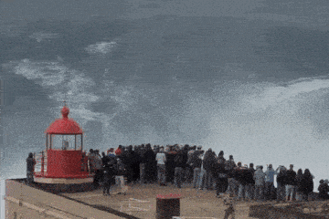 a group of people standing on top of a cliff looking at a large wave