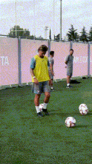a man in a yellow vest kicks a soccer ball in front of a pink wall that says ta