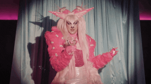 a drag queen in a pink outfit holds a glass of wine