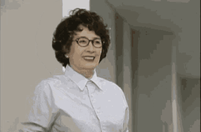 an elderly woman wearing glasses and a blue shirt is smiling while standing in a hallway .