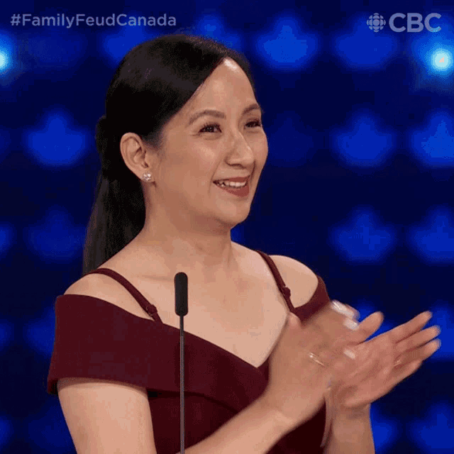 a woman in a red dress applauds in front of a microphone with the words family feud canada behind her