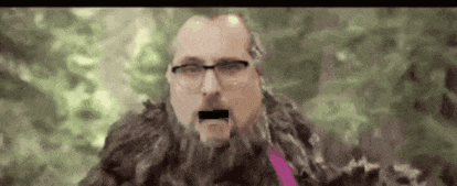 a man with glasses and a beard is standing in the woods .
