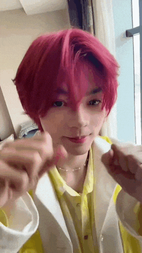 a young man with pink hair and a yellow shirt is making a heart with his hands .