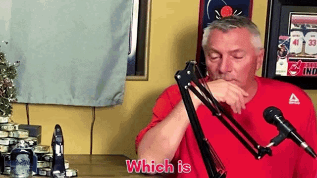 a man in a red shirt is sitting in front of a microphone with the words which is above him