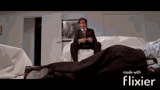 a man in a suit and tie is sitting on a couch next to a dead man with blood on his face