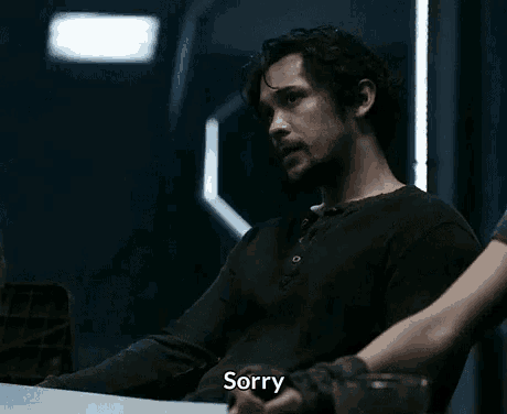 a man in a black shirt is sitting at a table with a person holding his hand and saying sorry .