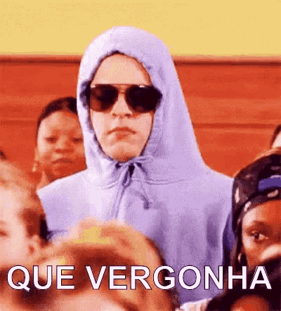 a man wearing a hoodie and sunglasses is standing in a crowd of people with the words que vergonha above him .