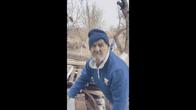 an old man wearing a blue sweater and a beanie is pointing his finger at the camera .