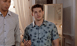 two men are standing next to each other in a room . one of the men is wearing a leafy shirt .